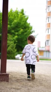 Beyaz bir erkek çocuk oyun parkındaki kumların yanından geçiyor. Küçük çocuk yazın dışarıda oynuyor. Dikey video.