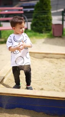 Küçük çocuk kum havuzunda oturuyor. Bebek ayağa kalkar ve ellerinden biraz kum döker. Yazın çocuk parkında. Dikey video.