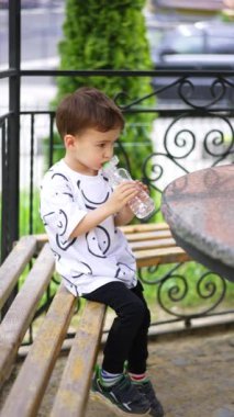 Beyaz çocuk bankta elinde şişeyle oturuyor. Çocuk biraz su içer ve şişeyi masaya koyar. Dikey video.