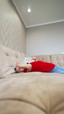 Neşeli erkek bebek kanepede yuvarlanıyor ve yere düşüyor. Mutlu çocuk halıda sürünüyor. Toddler evde oynuyor. Dikey video.