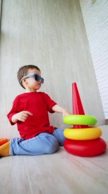 Kırmızı tişörtlü ve güneş gözlüklü bebek yerde oturuyor. Sevimli çocuk oyuncak piramidi monte ediyor. Düşük açı. Dikey video.