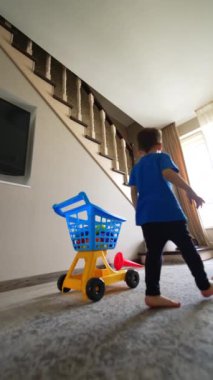 Küçük çocuk oyuncak arabasını odaya itiyor. Bebek merdivenlere basar ve ellerini sallar. Dikey video.