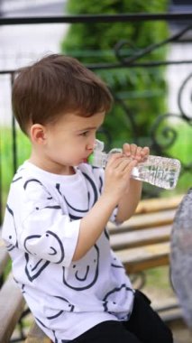 Beyaz tişörtlü, tatlı bir çocuk gülümseyerek şişeden su içiyor. Çocuk içkisini bitirir ve masaya bir şişe koyar. Dikey video.