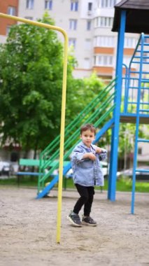 Kot pantolonlu bir çocuk oyun parkının yanından geçiyor. Küçük çocuk tişörtünü çıkarıp kaydırağa doğru gidiyor. Dikey video.