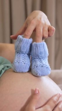 Minik bebek çoraplarıyla oynayan hamile bir kadın. Müstakbel anne evde dinlenirken mavi örgü çoraplarıyla göbeğine basıyor. Müstakbel anne hamileliğin tadını çıkarıyor. Dikey video