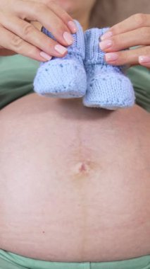 Hamile bir kızın karnı. Müstakbel anne çıplak karnında bebek çoraplarıyla oynuyor. Hamilelik sırasındaki sağlık kavramı. Yakın plan. Dikey video