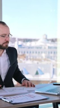 Ciddi bir ofis çalışanının portresi. Gözlüklü bir işadamı masasında oturuyor ve pencerenin yanındaki ofiste yeni bir proje üzerinde çalışıyor. Dikey video