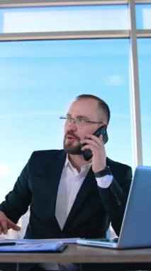 İş yerindeki girişimci portresi. Ofiste dizüstü bilgisayarın önünde otururken telefonla konuşan gözlüklü kendine güvenen bir iş adamı. Dikey video