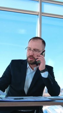 Ofiste çalışan ciddi bir iş adamı. Takım elbiseli sakallı patron masada dizüstü bilgisayarla oturup telefonla konuşuyor. İş konsepti. Dikey video