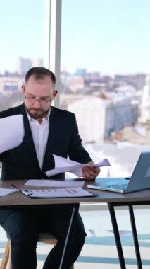 İş yerinde gergin bir girişimci var. Kostümlü bir iş adamı masada oturuyor ve kağıtları fırlatıyor. İş sorunları. Dikey video