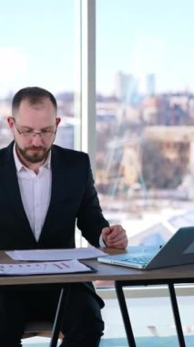 Takım elbiseli meşgul bir adam ofiste çalışıyor. Gözlüklü orta yaşlı girişimci bir sürü gazete ve dizüstü bilgisayarla masada oturuyor ve ciddi bir şekilde çalışıyor. Dikey video