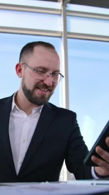 Gülümseyen iş adamının telefonundan bir video görüşmesi var. Gözlüklü, takım elbiseli, dizüstü bilgisayarlı, cep telefonu kullanan bir ofis çalışanı. Dikey video