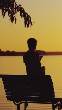 Küçük bir çocuk bankta oturuyor ve yazın gölün kenarında akşam manzarasının arka planında kağıt bir tekneyle oynuyor. Günbatımı Dikey video