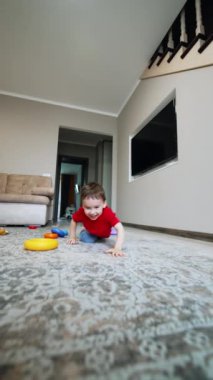Neşeli beyaz çocuk yerde sürünen kameraya doğru yaklaşıyor. Sevimli çocuk kameranın önünde yatıp bakıyor. Dikey video.