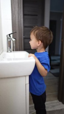 Güzel gülümseyen çocuk lavaboda durup suya bakıyor. Mutlu çocuk banyodaki musluğu kapatır. Dikey video.