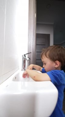 Mavi tişörtlü çocuk lavaboda ellerini yıkıyor. Küçük çocuk musluğu kapatır ve neşeli adımlarla yürür. Dikey video.