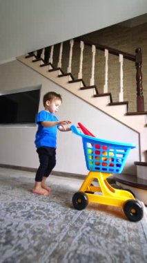 Mavi tişörtlü küçük çocuk arabasını oyuncaklarla birlikte alıyor. Küçük çocuk başka bir odaya tramvayı itiyor. Düşük açı. Dikey video.