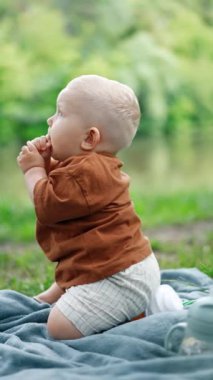 Kahverengi tişörtlü sevimli erkek bebek ekose üzerinde oturup bir şeyler yiyor. Yazın piknikte yürümeye başlayan çocuğun yan görüntüsü. Arkaplan bulanık. Dikey video.