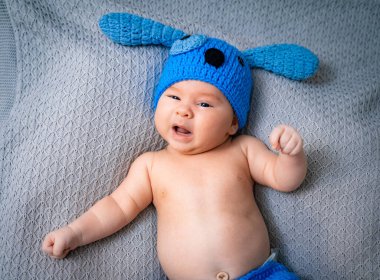 Mavi tavşan şapkalı bebek neşeli anın tadını çıkarıyor. Mavi tavşan şapkalı neşeli bir bebek, dokulu bir battaniyeye uzanmış mutluluk saçıyor..