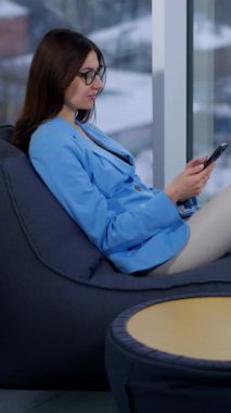 Esmer rahat ve rahat bir şekilde koltukta oturmuş telefonuna bakıyordu. Kadın haberlere kayıyor ya da internette sohbet ediyor ve sonra kameraya bakıyor. Arkaplan bulanık. Dikey video