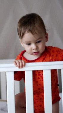 Turuncu takım elbiseli meraklı bebek karyolada dikiliyor. Küçük çocuk elinde bir emzik ile sevimli bir şekilde el sallamaya başlar. Dikey video