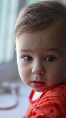 Pencerenin pervazında duran sevimli beyaz çocuk. Bebek kameraya doğru alaycı bir ifade takınıyor. Kapatın. Arkaplan bulanık. Dikey video