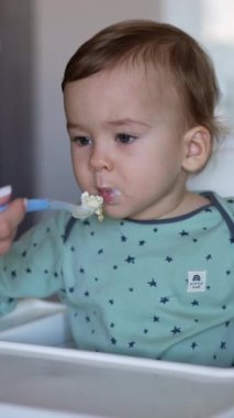 Bakıcı anne bebeğini kaşıkla besliyor. Annenin eli, beslenme sandalyesinde oturan çocuğuna kaşık veriyor. Arkaplan bulanık. Dikey video