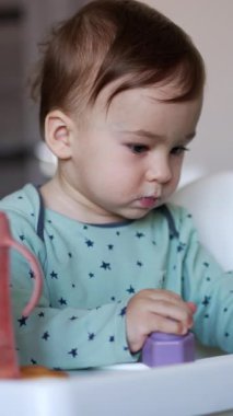 Beslenme koltuğunda oturan sevimli bebek. Yüzü lekelenmiş beyaz bir çocuk oyuncakla oynuyor. Arkaplan bulanık. Dikey video