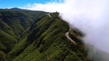 Yeşil çimlerle kaplı muhteşem yüksek dağlar. Kayanın tepesindeki otoyolda beyaz bulutlu bir yuvarlanma. Madeira, Portekiz. Hava görünümü.