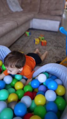 Sevimli neşeli çocuk plastik toplarla havuzda oturuyor. Çocuk iki sarı topu alıp el çırparak el arabasına koyuyor. Dikey video.
