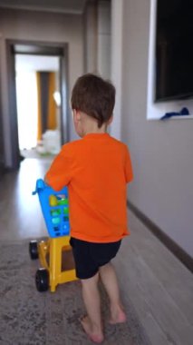 Koridorda yürüyen küçük bir çocuğu takip etmek arabayı itmek. Bebek evde mutlu bir şekilde oynuyor. Dikey video.