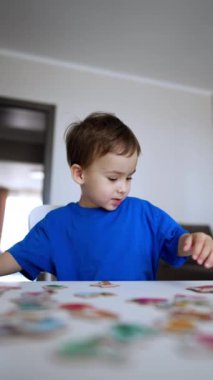 Mavi tişörtlü mutlu çocuk bir masaya koşuyor. Küçük çocuk masaya oturur ve bulmacaları inceler. Düşük açı. Dikey video.