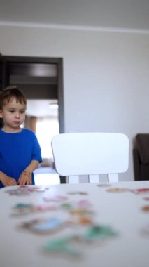 Genç bir çocuk masaya koşar ve sandalyeye oturur. Küçük çocuk bulmacalarla oynamaya başlar. Dikey video.