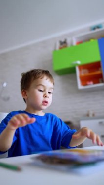 Mavi tişörtlü bebek boyama kitabının sayfalarını çeviriyor. Çocuk kalemleri masadan topluyor. Düşük açı. Dikey video.