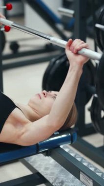 Genç, sıska bir kadın ayakları yere basan bir bankta uzanıyor ve spor salonunda iki koluyla ağırlık kaldırıyor. Yakın plan.