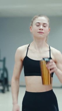 Siyah bluzlu güzel bir kadın sarı bir içecekten içiyor ve spor malzemelerinin arka planında spor salonunda yürüyor. Sağlıklı yaşam tarzı