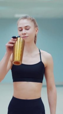 Siyah spor giysili genç bir kadın elinde bir sallayıcıyla spor salonuna giriyor ve ondan su içiyor. Spor salonu. Bulanık Arkaplan