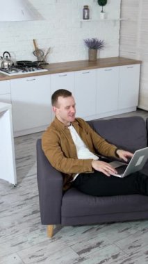 Evdeki kablosuz bilgisayarda çalışan rahat bir adam. Genç bir adam kanepeye uzanıyor ve modern mutfak arka planında dizüstü bilgisayar kullanıyor. Karantinada iş. Dikey video