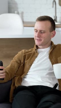 Evde telefon kullanan yakışıklı bir adam. Adam bir fincan kahveyle kanepede dinleniyor ve mutfaktaki akıllı bir telefonda fotoğraf çekiyor. Dikey video