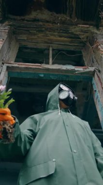 Mahvolmuş binada ekolojist. Koruyucu takım elbiseli ve bir demet çiçekli gaz maskeli biri. Kimyasal bölgede hava kirliliği. Aşağıdan bak. Dikey video