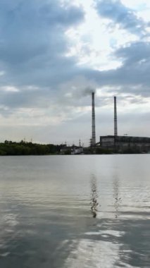 Endüstriyel bölgenin arka planında büyük borularla akşam suyu. Mavi gökyüzünün altındaki fabrika borularından beyaz duman dökülüyor. Çevre kirliliği. Dikey video