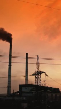 Kırmızı günbatımında zararlı fabrikanın yanındaki elektrik direkleri. Kara duman endüstriyel borudan havaya yayıldı. İletişim hatları. Çevreyi kirletiyor. Kamera aşağı iniyor. Dikey video