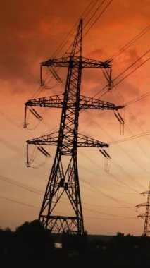 Günbatımında iletim hatları. Kızıl gökyüzüne karşı yüksek voltajlı elektrik desteği silueti. Elektrik direkleri yakınındaki endüstriyel borulardan gelen siyah duman havaya karışıyor. Dikey video