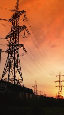 Günbatımında yüksek voltaj kablosu ve pilonlar. Kırmızı batan güneşli dramatik gökyüzüne karşı elektrik kulelerinin siluetleri. Dikey video