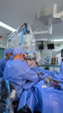 İki doktor ameliyat masasında oturmuş mikroskoba bakıyor. Tanınmayan hemşire cerrahi standında duruyor ve aletleri ayarlıyor. Dikey video.