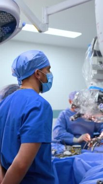 Şapkalı, maskeli ve üniformalı bir erkek doktor ileriye bakıyor. Cerrahlar mikroskop kullanarak arka planda ameliyat yaparlar. Dikey video.