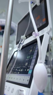 Serum ve tuz solüsyonu monitörlerin yanında asılı. Arka planda akciğer havalandırma makinası ekranları yaşam sinyallerini gösteriyor. Dikey video.