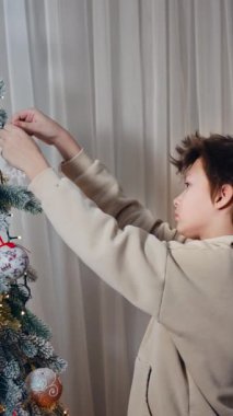 Evde güzel bir Noel ağacı süsü var. Genç çocuk bir gülümseme ile oyuncak kar tanesine bakar ve sonra onu ağaca koyar. Dikey video