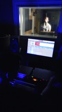 Karanlık profesyonel müzik stüdyosunda çalışan ses mühendisi. Şarkıcı yan pencerenin arkasında gösteri yapıyor. Dikey video