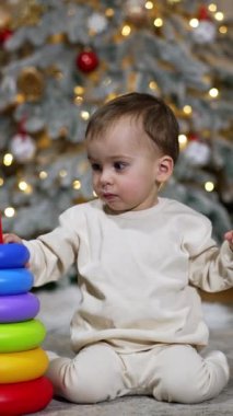 Güzel beyaz bebek oyuncak piramit inşa ediyor. Bebek, Noel ağacından çok uzak olmayan bir yerde yerde oturuyor. Dikey video